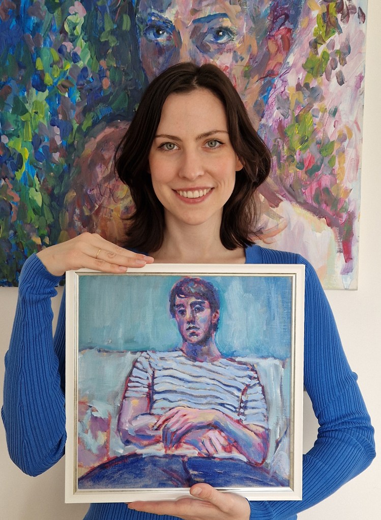 Artist Clara Niniewski holding in her hands the framed painting that will be exhibited at the New English Art Club (NEAC) annual exhibition at the Mall Galleries in London in June 2023. The painting is a portrait of a seated man, with a blue background and lots of blues and pink in the skin tone.